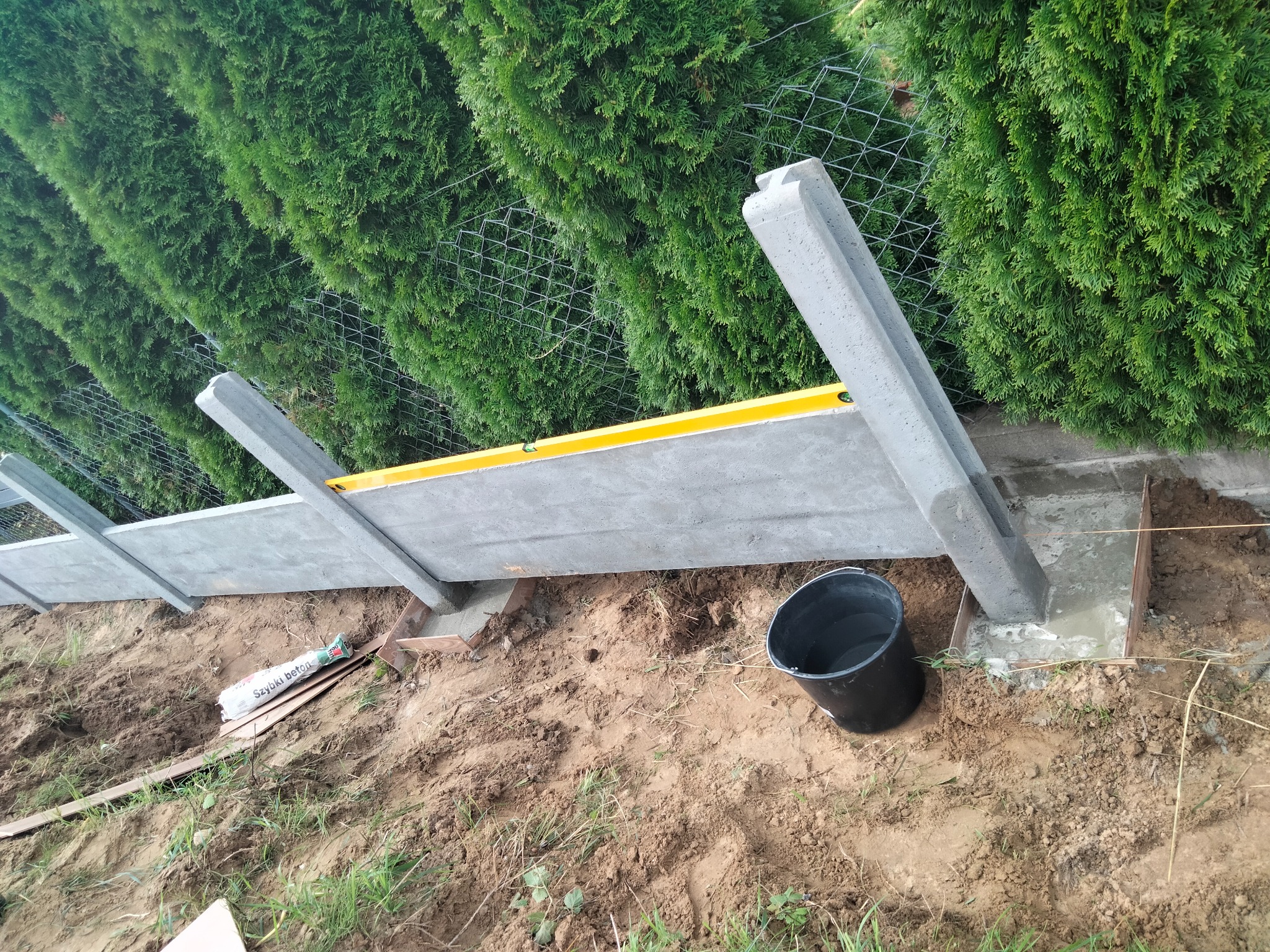 W Gryfinie trwa montaż betonowego ogrodzenia: widoczne słupki i panele w trakcie poziomowania, w tle zielony żywopłot i siatka.