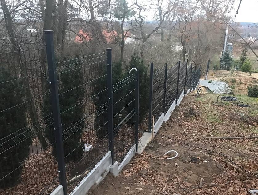 Czarny ogrodzenie panelowe na betonowej podmurówce, widok z góry na nierówny teren z drzewami w tle, kabel elektryczny leżący na ziemi.