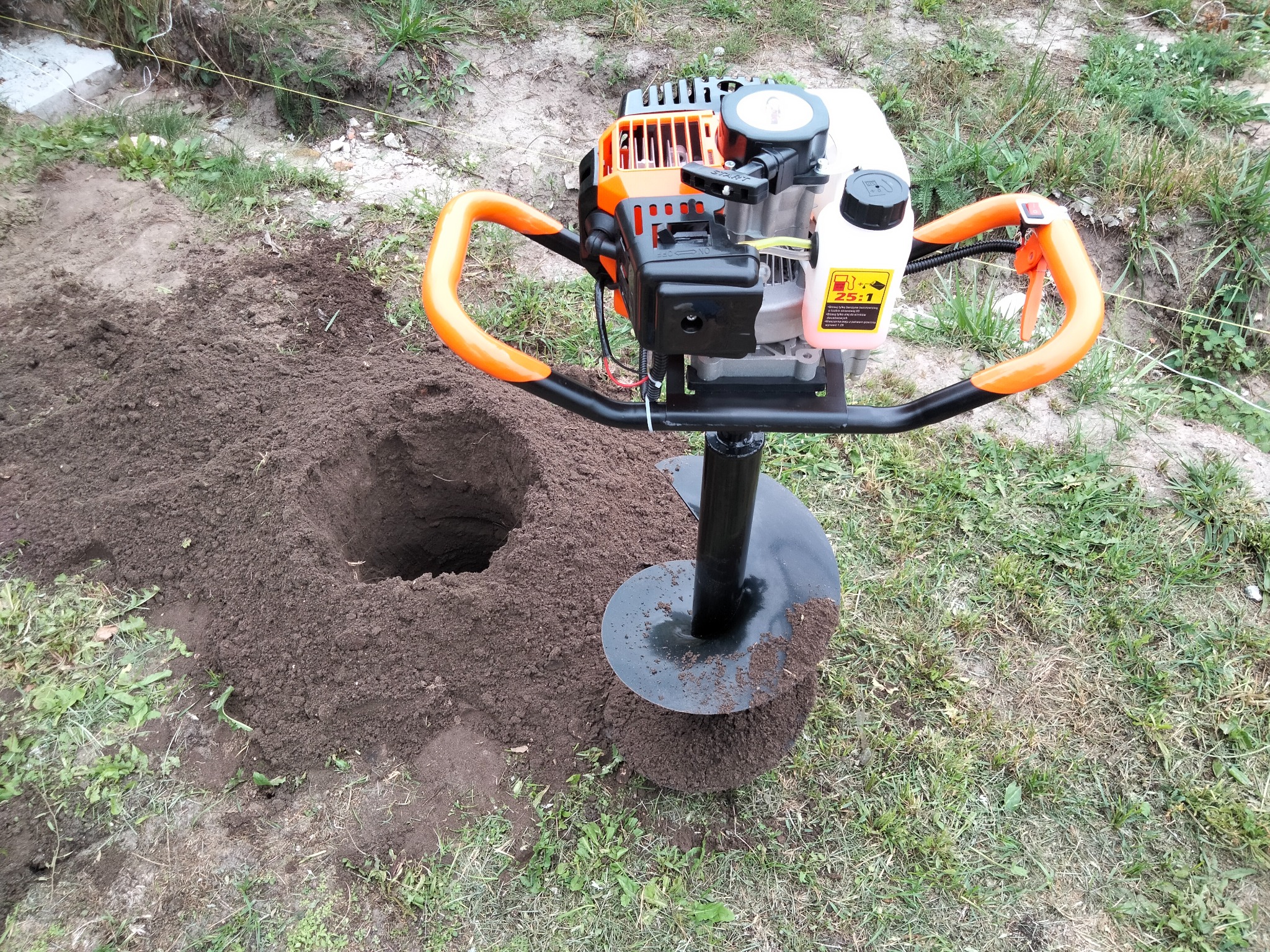 Wiertnica spalinowa do gruntu z pomarańczowymi uchwytami, obok wykopany otwór w ziemi, trawa w tle.