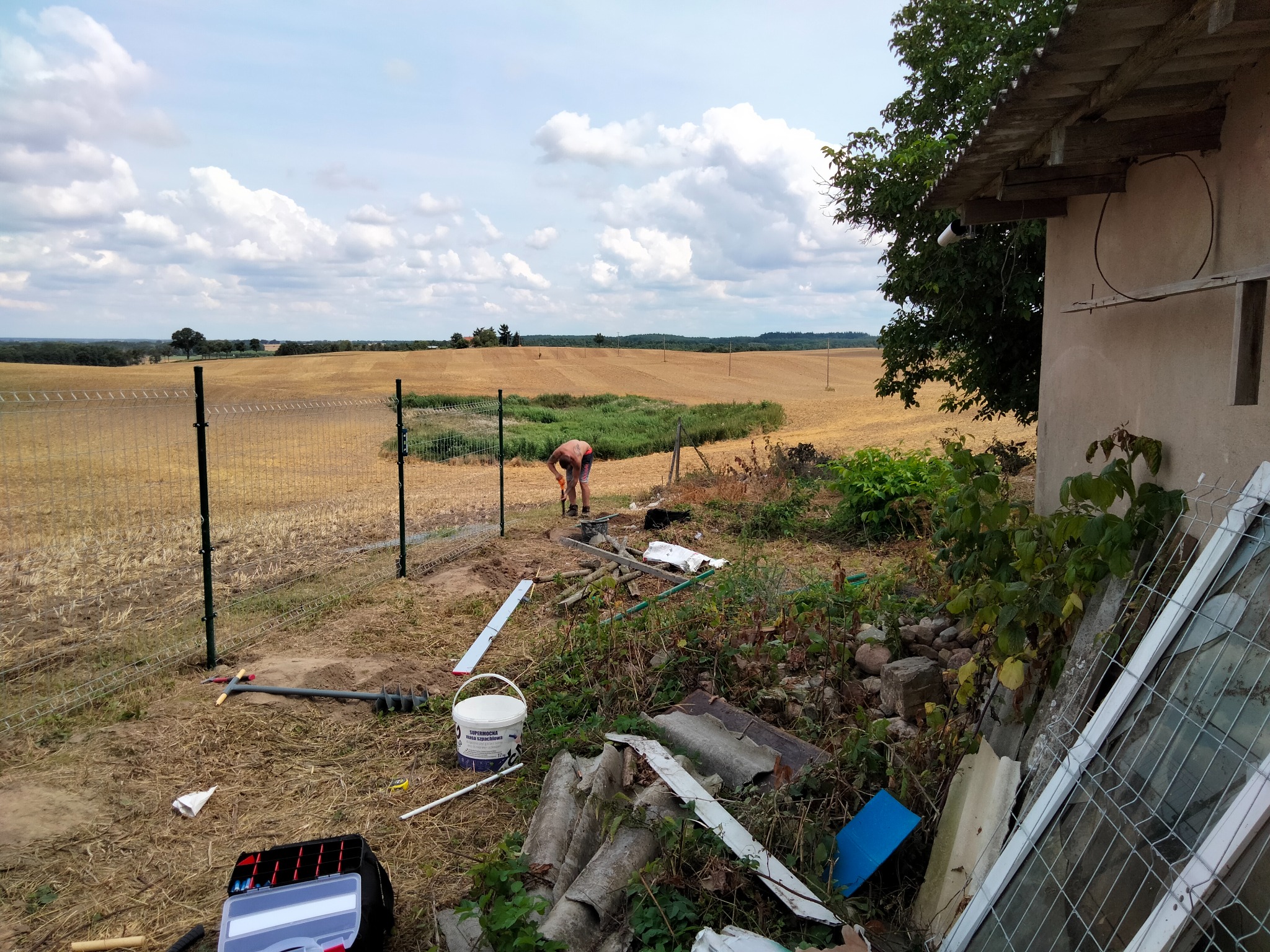 Montaż ogrodzenia panelowego na polu, widoczny pracownik w trakcie pracy, narzędzia i materiały budowlane w pobliżu, krajobraz polny w tle.