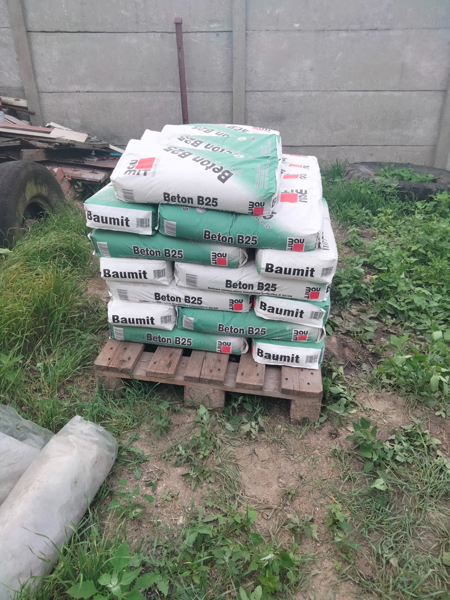 Stos worków z betonem Baumit B25 na drewnianej palecie, ustawiony na ziemi z trawą, z betonowym ogrodzeniem w tle.