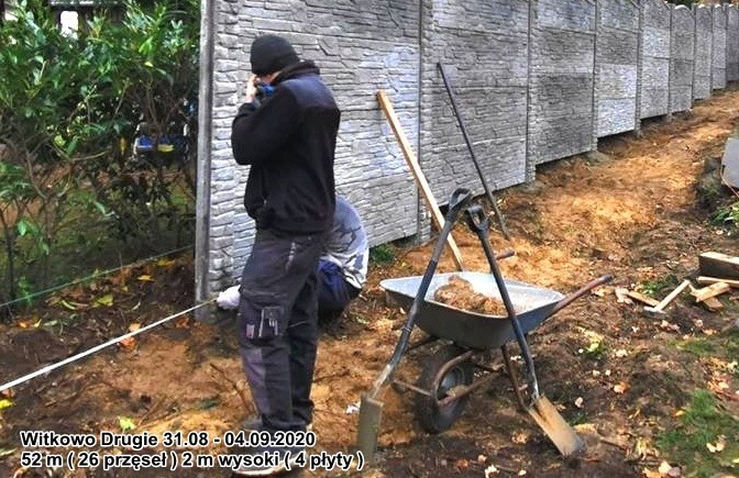 Montaż betonowego ogrodzenia imitującego kamień; widoczni dwaj pracownicy, jeden przy słupku, drugi w kucki, taczka z zaprawą i narzędzia na ziemi.