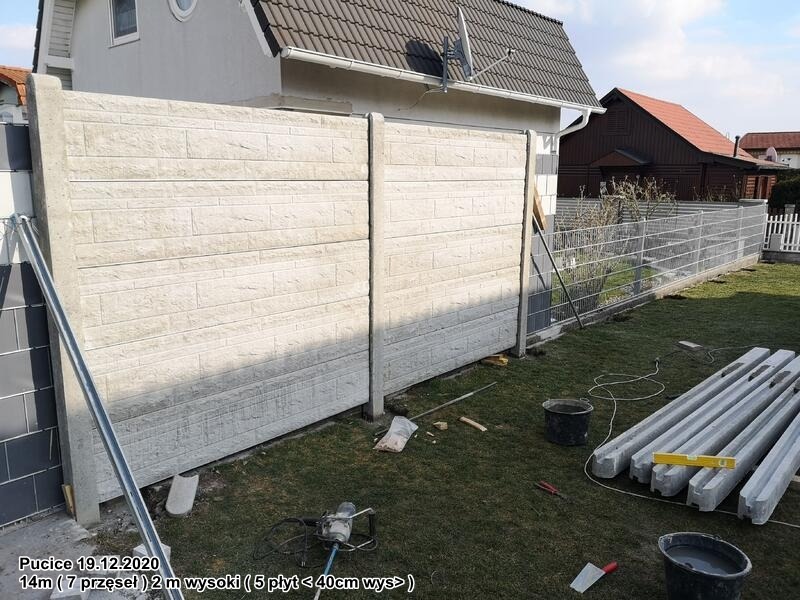 Montaż ogrodzenia betonowego z prefabrykatów: widoczne dwa panele z fakturą cegły, słupki, przygotowane elementy, narzędzia i wiaderka z zaprawą na trawniku, w tle dom i ogrodzenie z siatki.