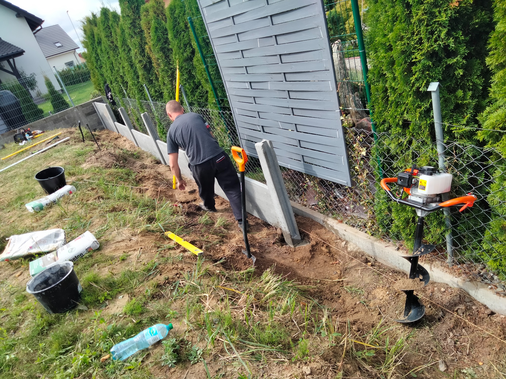 Mężczyzna w trakcie montażu betonowego ogrodzenia, używa poziomicy do ustawienia słupka, w tle widoczny świder do wiercenia otworów w ziemi i częściowo zamontowane panele ogrodzeniowe z szarego...