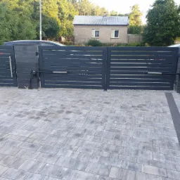 Nowoczesna, automatyczna brama wjazdowa z poziomymi panelami w kolorze antracytowym, zamontowana na grafitowych słupkach z bloczków betonowych, widok z zewnątrz na podjazd wyłożony szarą kostką...