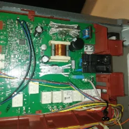 Otwarta obudowa urządzenia AGD z widoczną, zieloną płytą drukowaną, elementami elektronicznymi i okablowaniem. Wnętrze urządzenia o czerwono-brązowej ramie. Widoczne kondensatory, transformator...