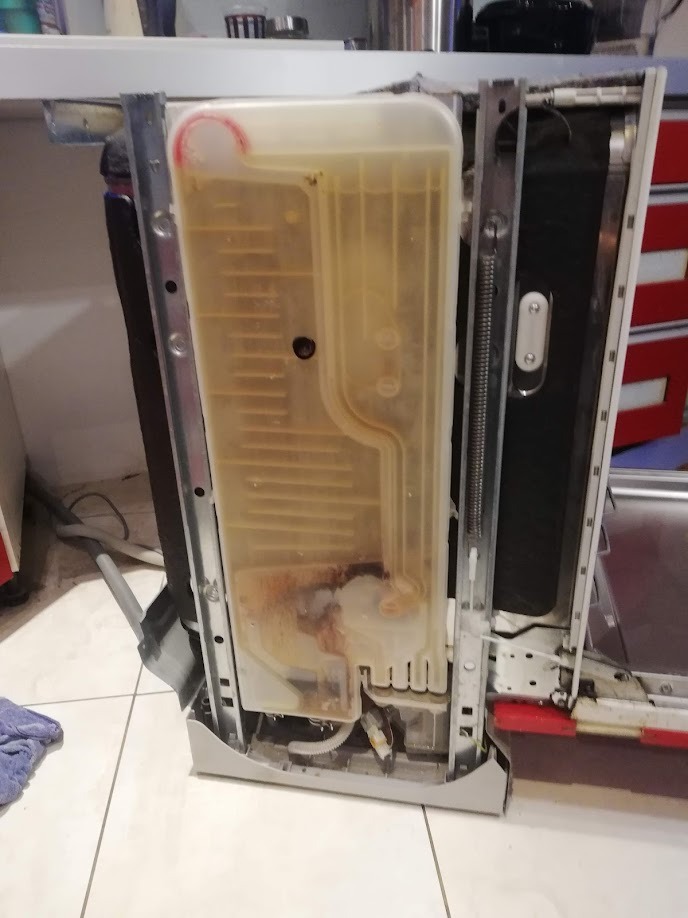 Wnętrze zdemontowanej zmywarki z widocznym, zabrudzonym pojemnikiem na sól i elementami metalowymi. Widoczne ślady użytkowania i osady.