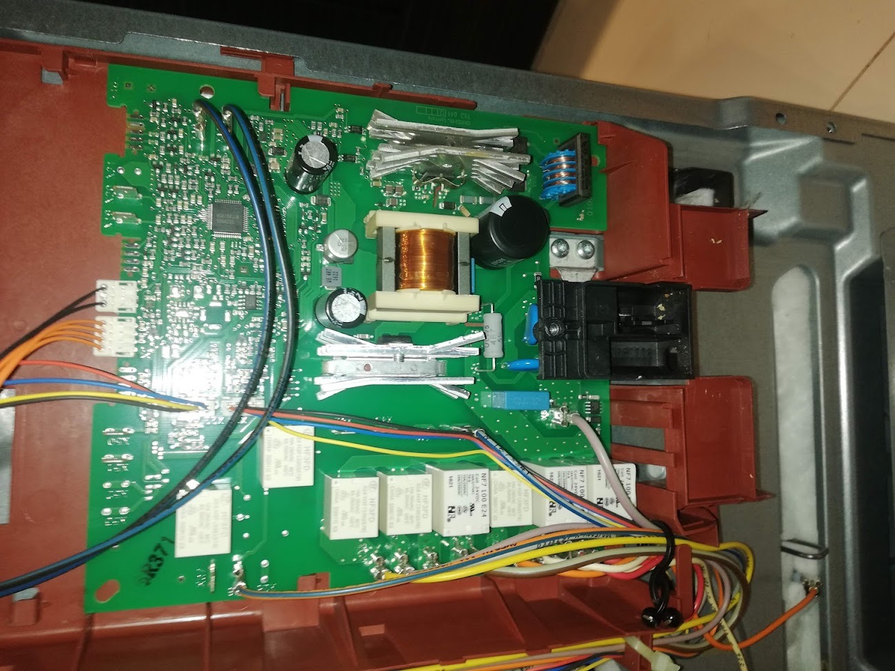 Otwarta obudowa urządzenia AGD z widoczną, zieloną płytą drukowaną, elementami elektronicznymi i okablowaniem. Wnętrze urządzenia o czerwono-brązowej ramie. Widoczne kondensatory, transformator...