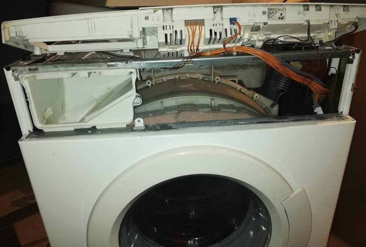 Otwarta pralka automatyczna podczas naprawy, widoczne wnętrze z przewodami i elementami konstrukcyjnymi, górna część obudowy uniesiona.