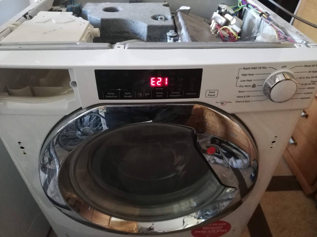 Otwarta pralka z widocznym błędem E21 na wyświetlaczu, panel sterowania z różnymi programami prania, wnętrze częściowo zdemontowane, widoczne przewody i elementy konstrukcyjne.