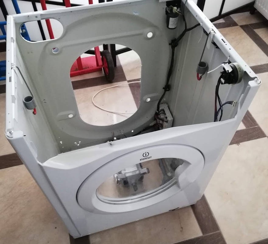 Rozłożona pralka marki Indesit z widocznymi elementami wewnętrznymi i drzwiczkami, na tle podłogi z płytek.