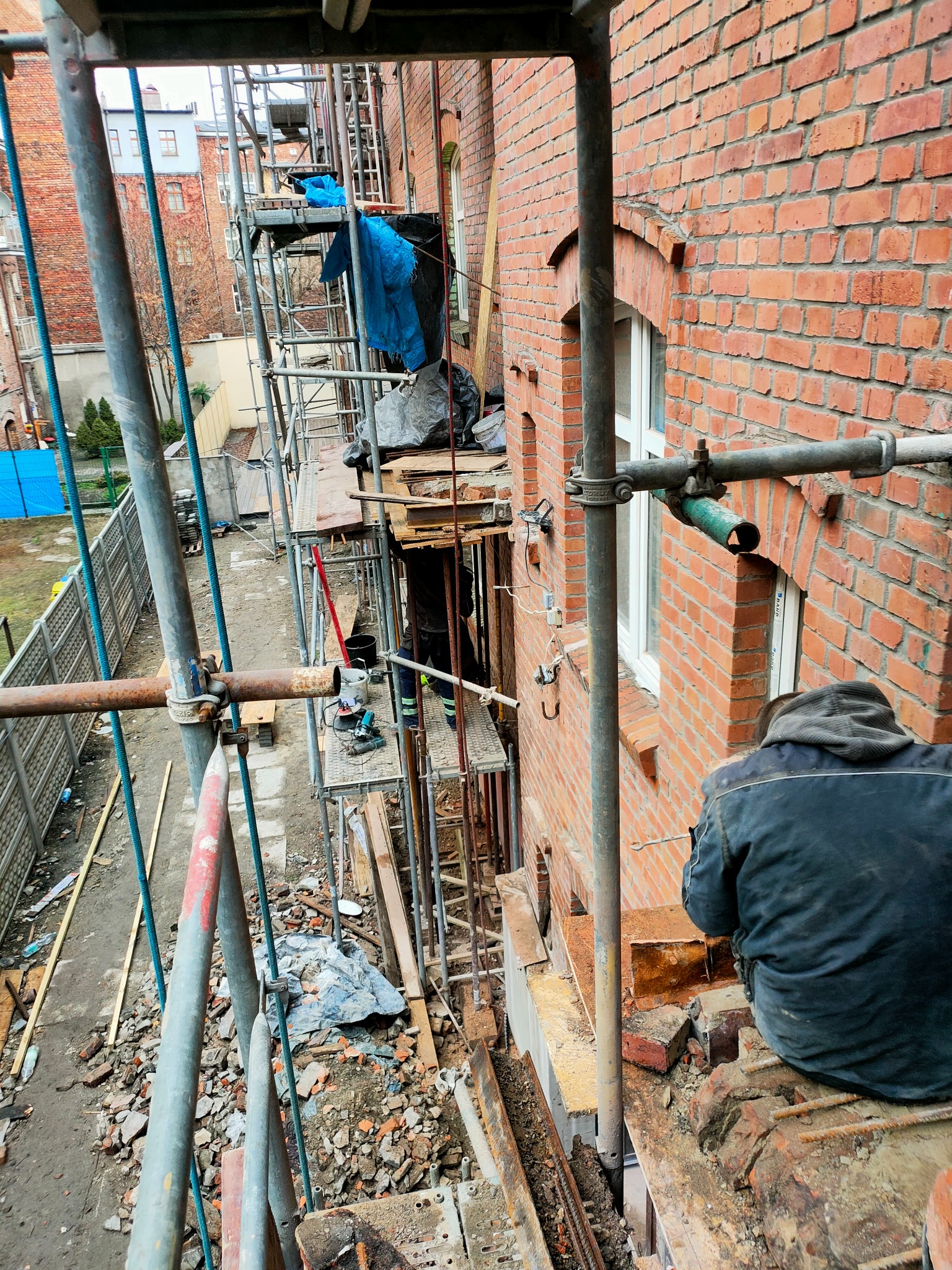 Murarz siedzi na rusztowaniu przy ceglanym budynku, widoczne uszkodzenia muru i przygotowania do prac remontowych.