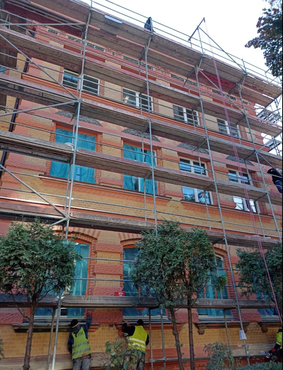 Budynek w rusztowaniach z zabezpieczonymi oknami, pracownicy w kamizelkach wykonują prace na elewacji.