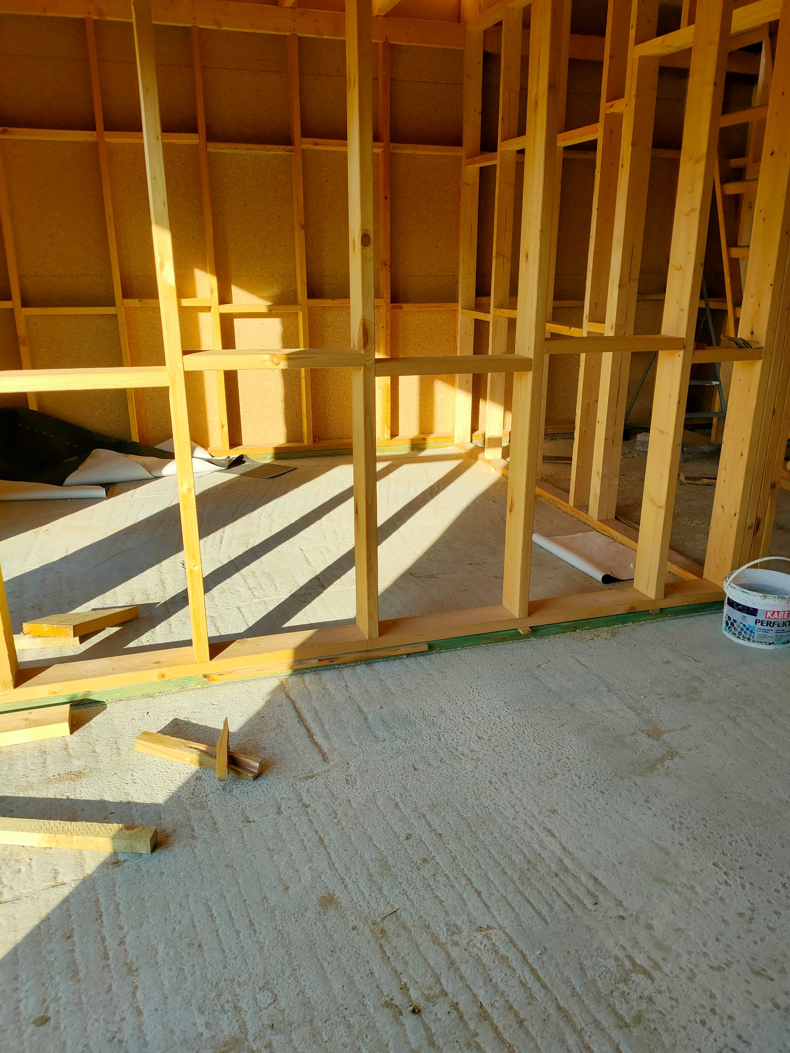 Surowe drewniane ramy ścian wewnętrznych w trakcie budowy, z widocznym betonowym podłożem, przygotowane do dalszych prac wykończeniowych, oświetlone naturalnym światłem słonecznym wpadającym przez...