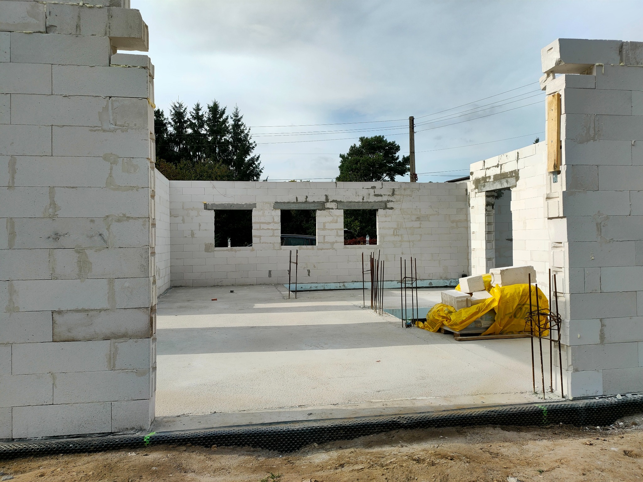 Wznoszenie ścian domu z bloczków betonu komórkowego, widoczne otwory okienne i drzwiowe, zbrojenie pionowe, paleta z bloczkami zabezpieczona żółtą folią.