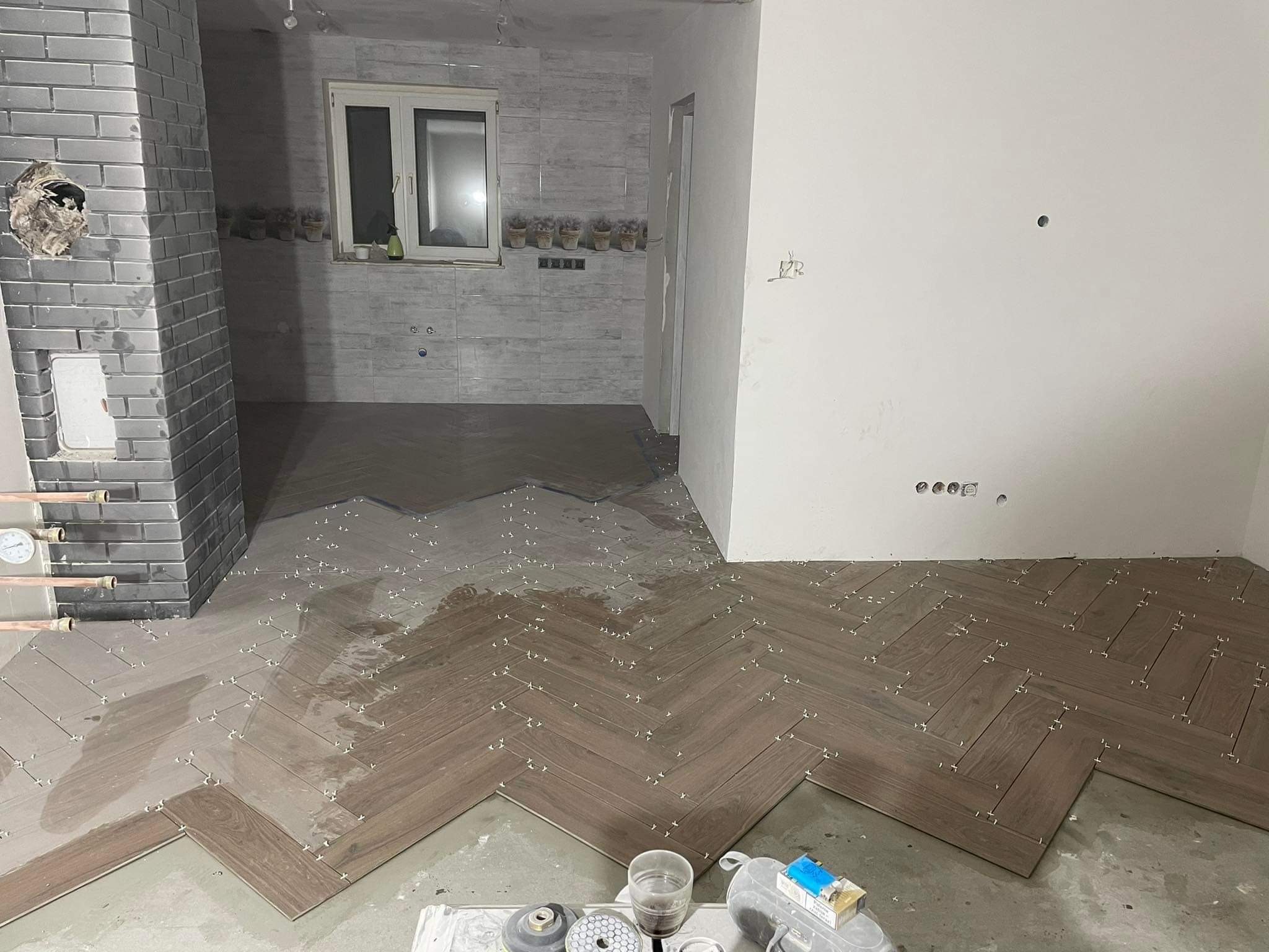 Układanie parkietu w jodełkę w trakcie remontu, widoczne płytki, krzyżyki dystansowe, narzędzia glazurnicze oraz fragment komina z szarej cegły.