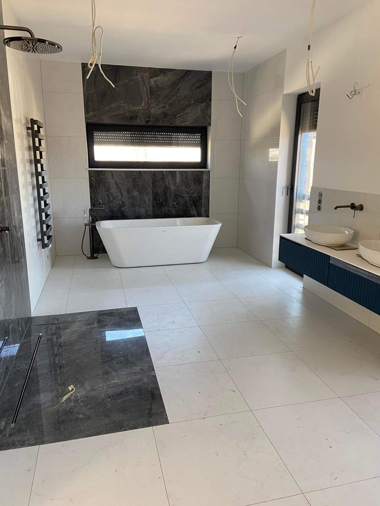 Nowoczesna łazienka w trakcie wykańczania: białe i szare płytki, wolnostojąca wanna z czarną armaturą, wisząca szafka z umywalkami, widoczne przewody elektryczne.