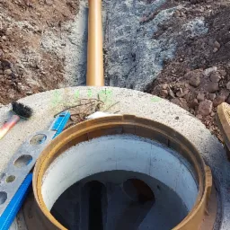 Betonowy krąg studni z otwartym włazem, widoczny rurociąg w wykopie, poziomica i szczotka na krawędzi, w tle koparka.