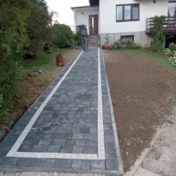 Ułożona ścieżka z szarej kostki brukowej z białymi obramowaniami prowadząca do domu, widoczna osoba w oddali oraz przygotowany teren pod trawnik.