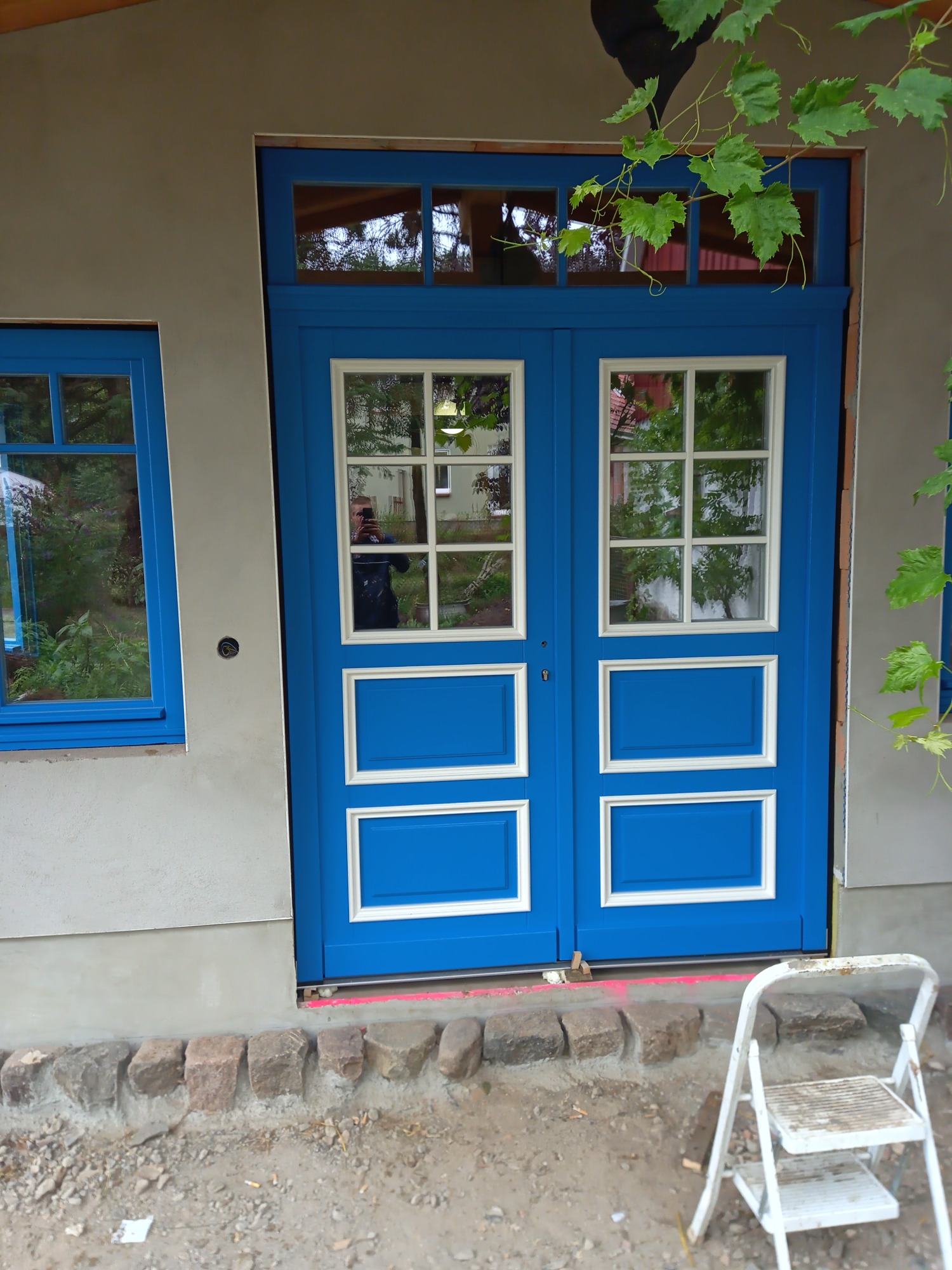 Niebieskie, dwuskrzydłowe drzwi wejściowe z białymi ramkami i szybami, widoczne podczas montażu, z drabinką opartą o próg i oknem obok, na tle surowej elewacji z widocznymi śladami prac budowlanych.