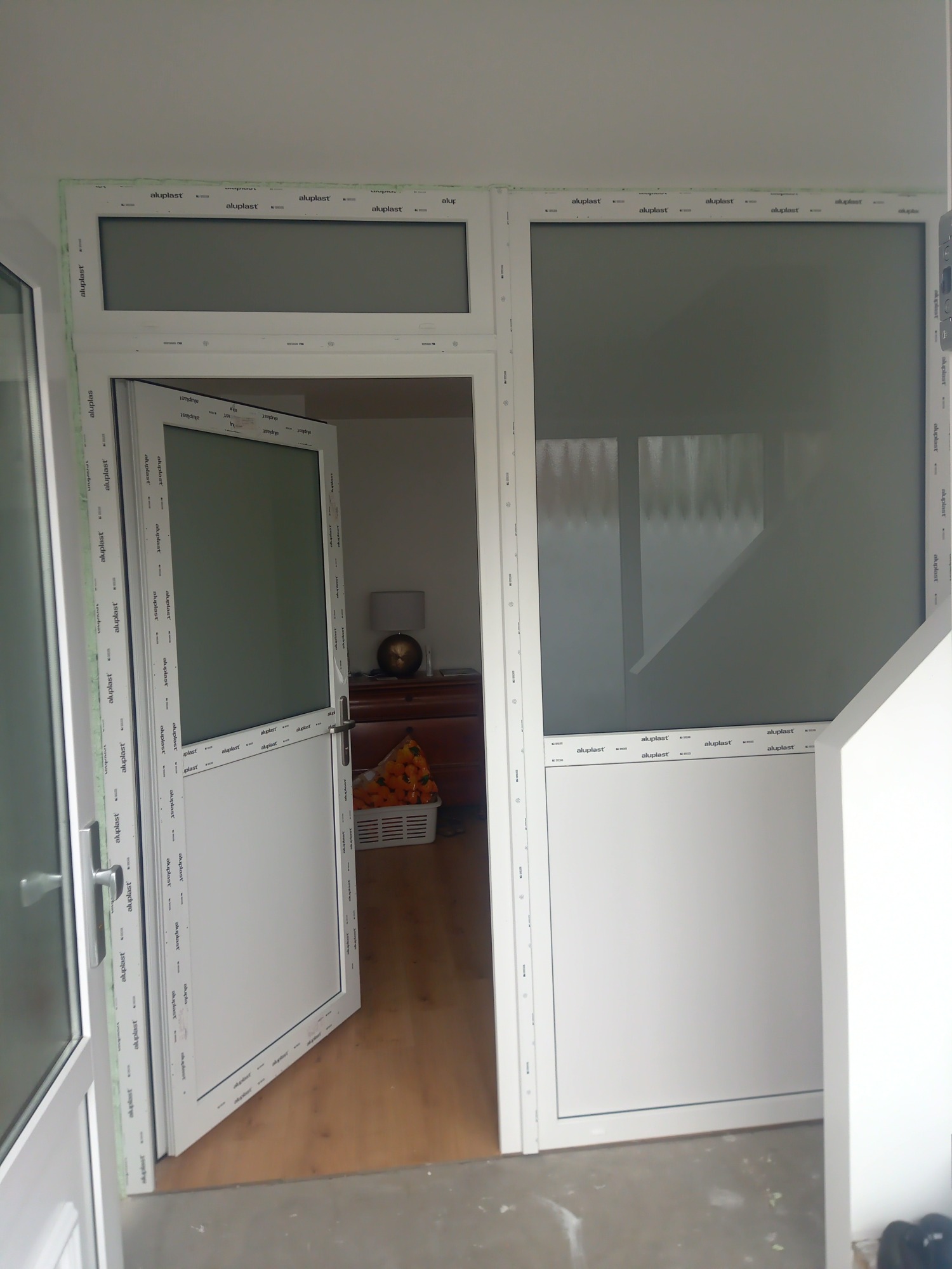 Białe, przeszklone drzwi balkonowe z ramą z tworzywa sztucznego, otwarte do wnętrza, widok na pokój z komodą i lampką nocną, widoczne ślady montażu na podłodze.