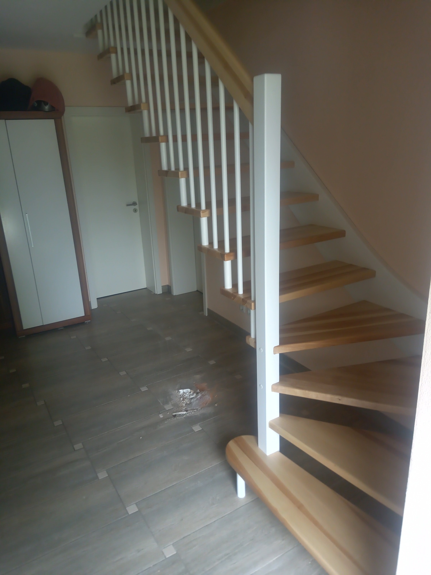 Jasne drewniane schody z białymi balustradami w jasnym wnętrzu, widoczne płytki na podłodze i uszkodzenie w podłodze.