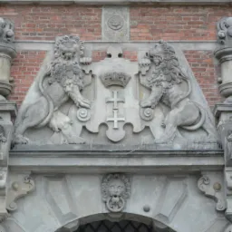 Kamienna fasada budynku z dwoma lwami podtrzymującymi herb z krzyżem i koroną, zdobiona rzeźbami twarzy po bokach, na tle ceglanej ściany.