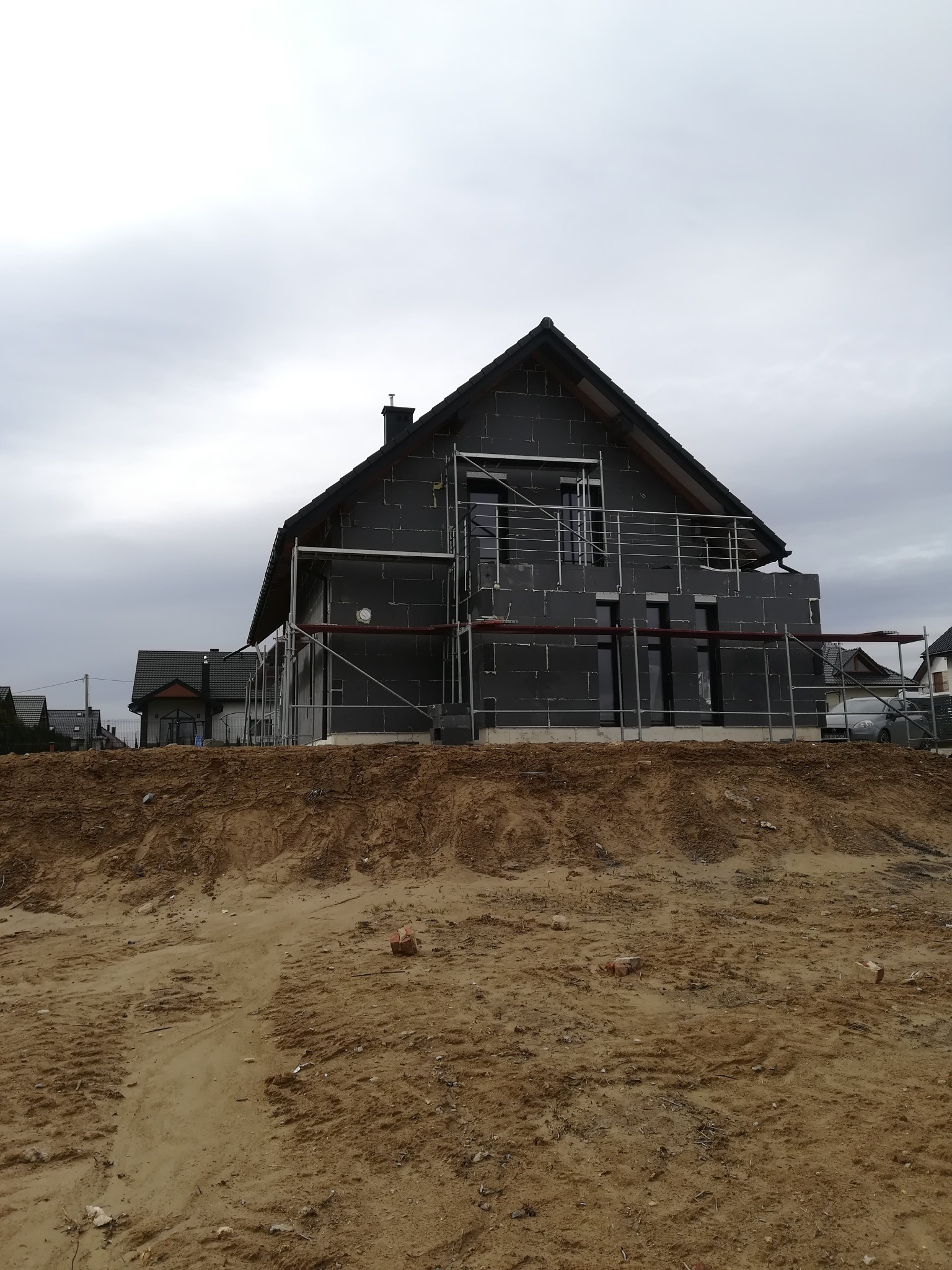 Dom jednorodzinny w trakcie ocieplania styropianem grafitowym, widoczne rusztowania i wykop ziemny, pochmurne niebo.