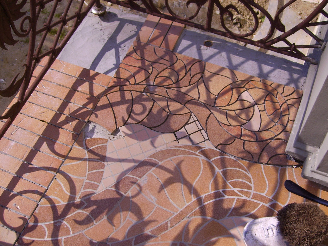 Układanie mozaiki ceramicznej o wzorze roślinnym na tarasie, widoczne narzędzia glazurnicze i cień ozdobnej balustrady.