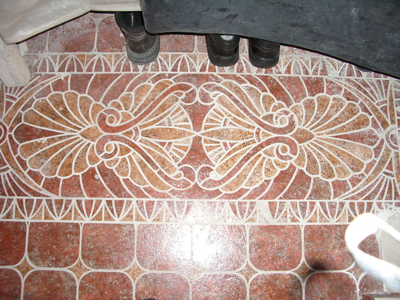 Wzorzysta, brązowo-biała posadzka z płytek ceramicznych w łazience, widoczne fragmenty wanny i stóp osoby wykonującej prace.