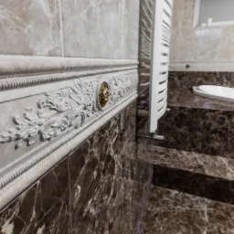 Elegancka łazienka z marmurowymi płytkami w odcieniach beżu i brązu, ozdobnym gzymsem z reliefami i złotym akcentem, oraz białym grzejnikiem drabinkowym.