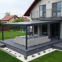 Zadaszony taras z szarą podłogą kompozytową, oświetleniem punktowym i białymi kamykami dekoracyjnymi, przylegający do domu z balkonem i zielonym trawnikiem w tle.