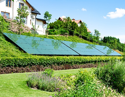 Instalacja paneli słonecznych na zboczu wzgórza, widoczne domy w tle, trawnik i roślinność.