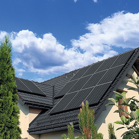 Czarny dach skośny z panelami słonecznymi w układzie prostokątnym, błękitne niebo z chmurami w tle, widok z poziomu gruntu, iglaki w pierwszym planie.