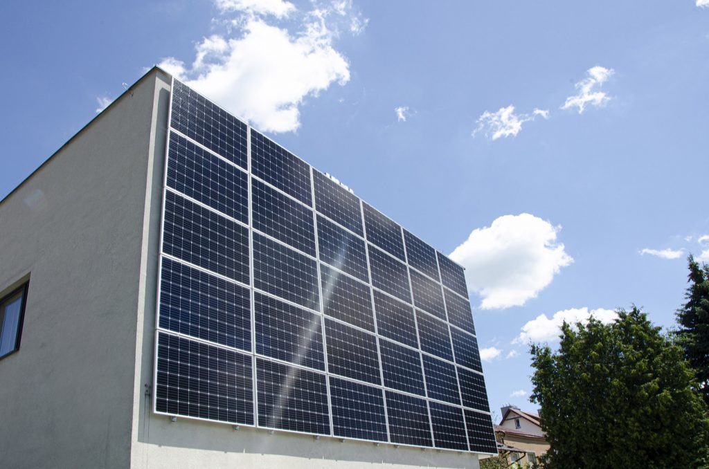 Duży panel fotowoltaiczny na ścianie domu jednorodzinnego, widok z dołu, błękitne niebo z obłokami w tle.