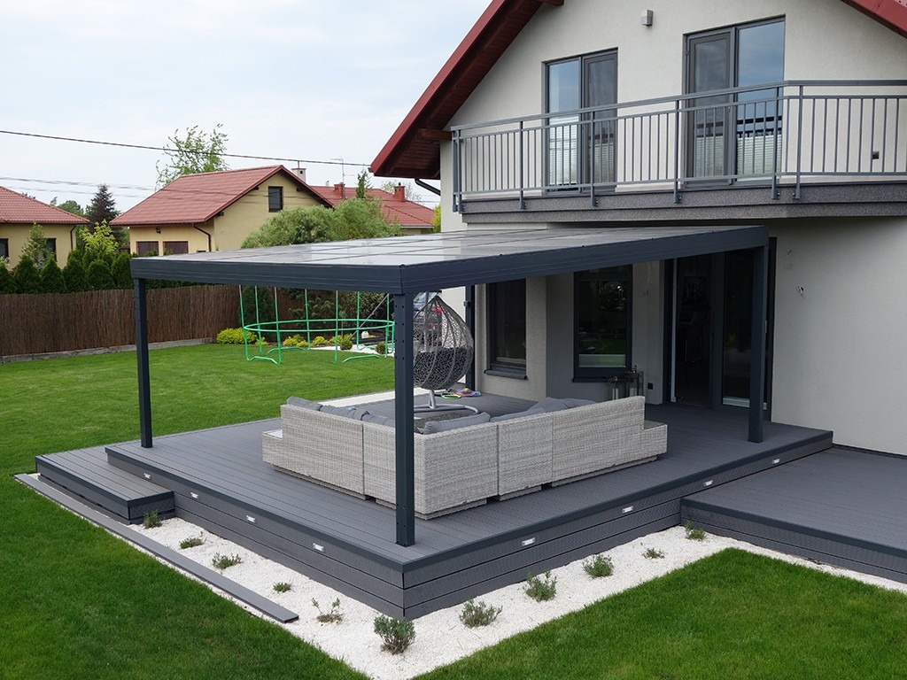 Zadaszony taras z szarą podłogą kompozytową, oświetleniem punktowym i białymi kamykami dekoracyjnymi, przylegający do domu z balkonem i zielonym trawnikiem w tle.