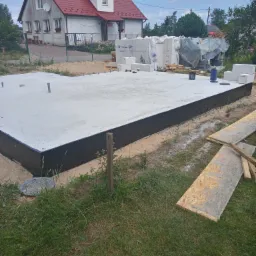 Wylana płyta fundamentowa z czarną izolacją, widoczne wystające rury instalacyjne, bloczki fundamentowe i dom w tle.
