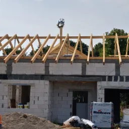 Wznoszenie domu murowanego z betonu komórkowego, widoczna konstrukcja dachu z wieńcem, materiały budowlane Roben na pierwszym planie, piasek i puszki z farbą w otworze okiennym.