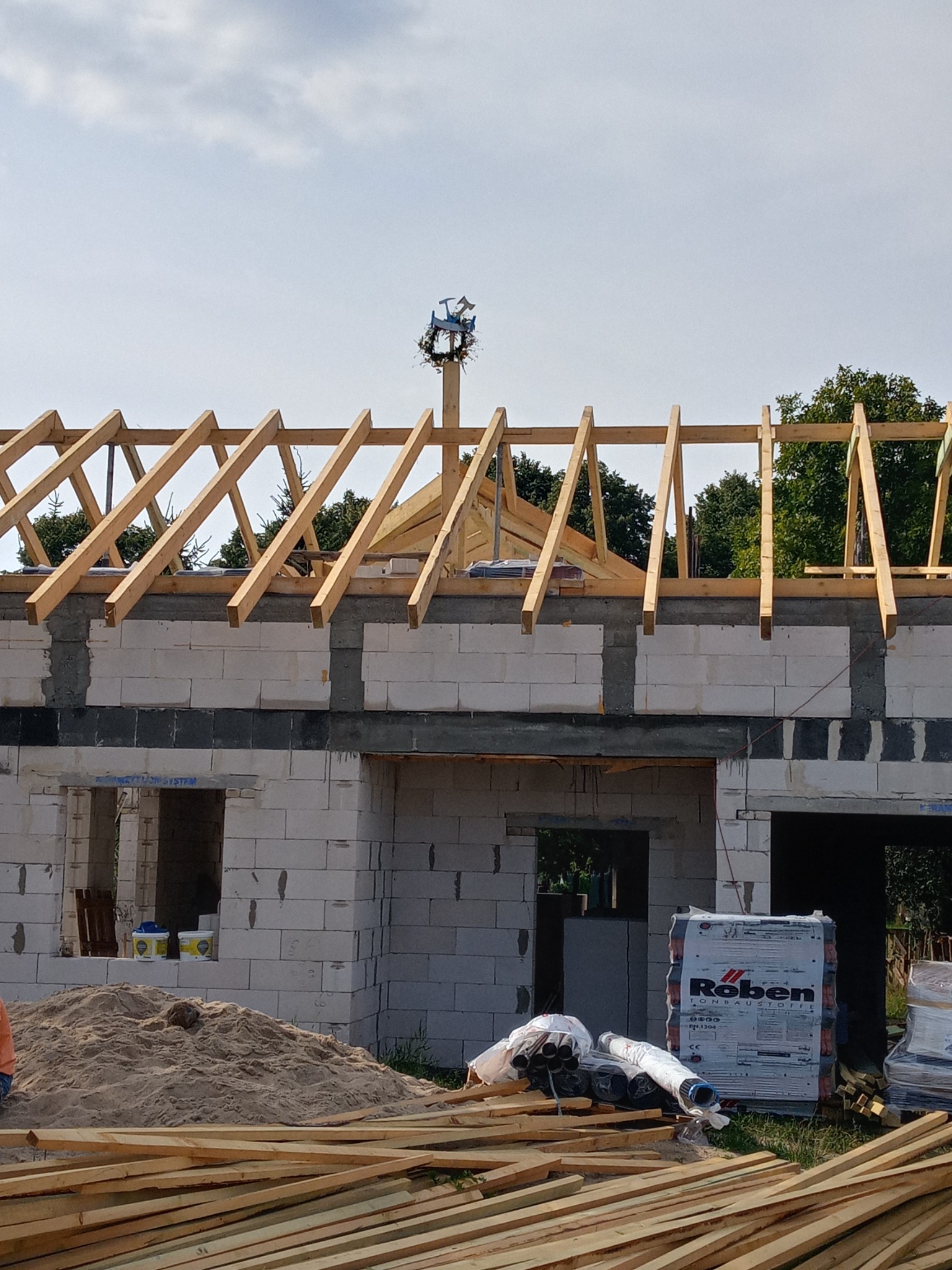 Wznoszenie domu murowanego z betonu komórkowego, widoczna konstrukcja dachu z wieńcem, materiały budowlane Roben na pierwszym planie, piasek i puszki z farbą w otworze okiennym.