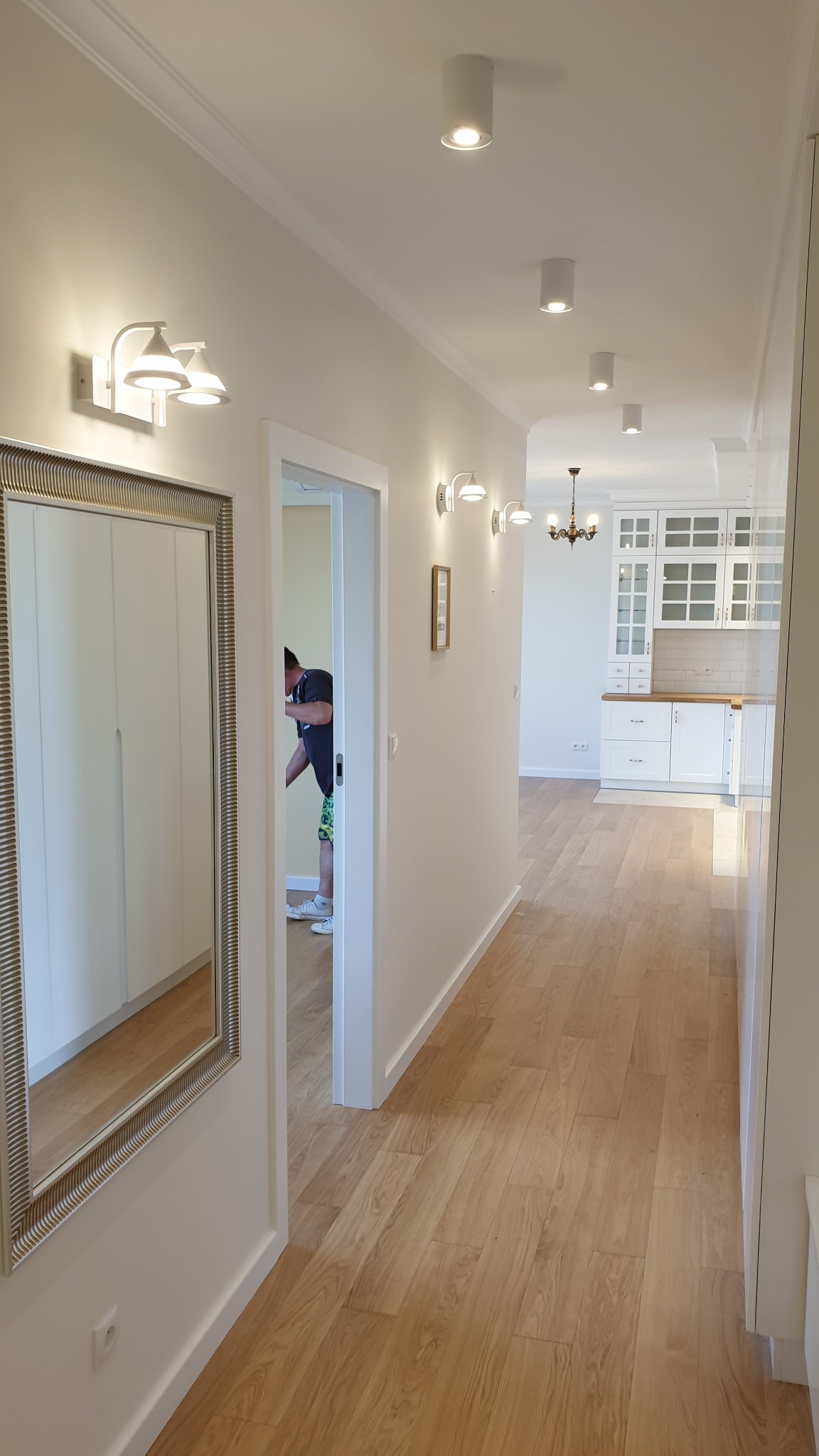 Korytarz w jasnych barwach z drewnianą podłogą, oświetleniem punktowym i kinkietami, widoczne lustro w ozdobnej ramie i częściowo widoczna biała kuchnia w tle, osoba w przejściu.