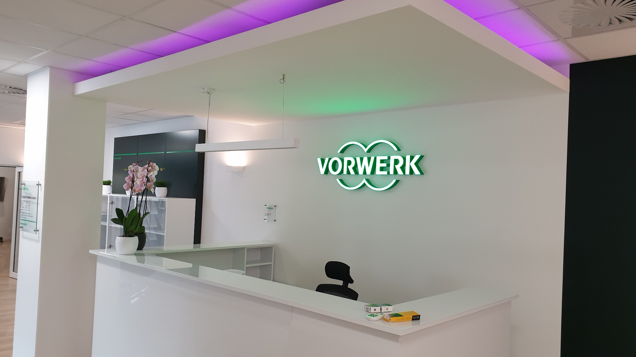 Nowoczesne biuro recepcyjne z białymi ścianami, podświetlanym logo firmy Vorwerk w kolorze zielonym, roślinami w doniczkach i elementami oświetlenia LED w kolorze fioletowym.