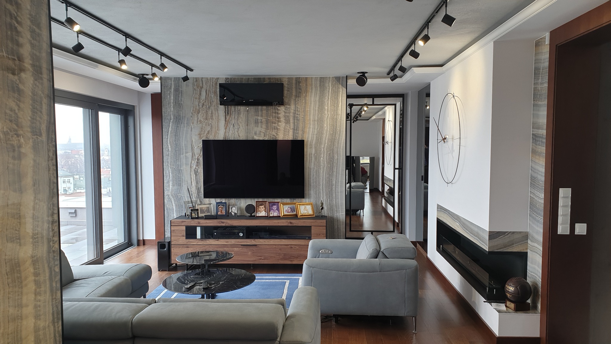 Nowoczesny salon z szarą sofą, ścianą z kamienia dekoracyjnego, telewizorem, podwieszanym oświetleniem punktowym i kominkiem z dekoracyjnym zegarem na ścianie.