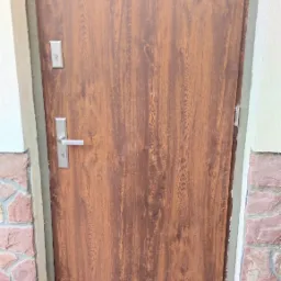 Brązowe drzwi wejściowe z imitacją drewna, zamontowane w ościeżnicy z widocznym uszczelnieniem, na tle kamiennej elewacji i kostki brukowej.