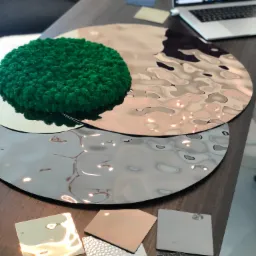 Pr&oacute;bki materiał&oacute;w wykończeniowych: metalowe krążki z fakturą, kwadratowe płytki w r&oacute;żnych odcieniach i fakturach, zielony okrągły element dekoracyjny na tle laptopa z projektem wnętrza.