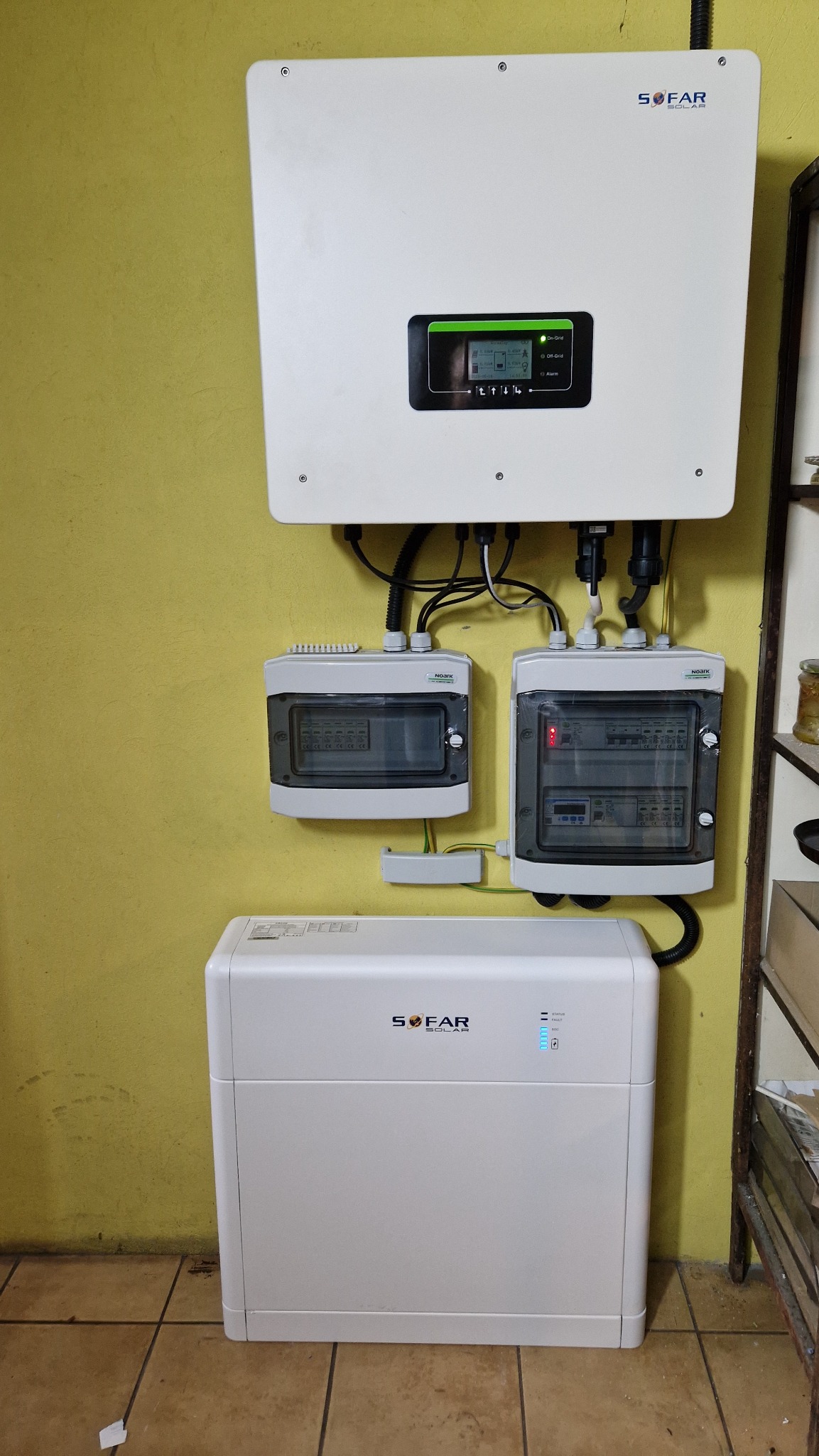 Biały falownik SOFAR Solar z wyświetlaczem, dwoma rozdzielnicami i magazynem energii na żółtej ścianie wewnątrz pomieszczenia.