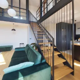 Nowoczesne wnętrze z drewnianymi schodami na metalowej konstrukcji, widoczne z dołu, obok zielona sofa i pufa w salonie, w tle przeszklona antresola i fragment kuchni.