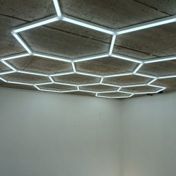 TK EL Instalacje Elektryczne TOMASZ KOWALSKI - Geometryczna instalacja oświetleniowa LED w kształcie plastra miodu na surowym betonowym suficie. Nowoczesny design, jasne światło. Widok z dołu na białe ściany.