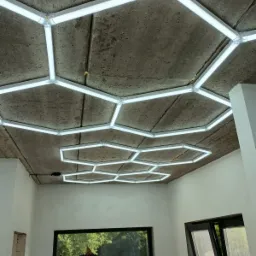 Geometryczna instalacja oświetleniowa LED na surowym betonowym suficie w pomieszczeniu z oknami i drabiną, widok z dołu.