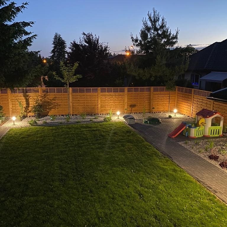 Oświetlony ogród z drewnianym ogrodzeniem, ścieżką z kostki i lampami. Widoczne elementy placu zabaw i robot koszący. Ujęcie wieczorne, z delikatnym oświetleniem.