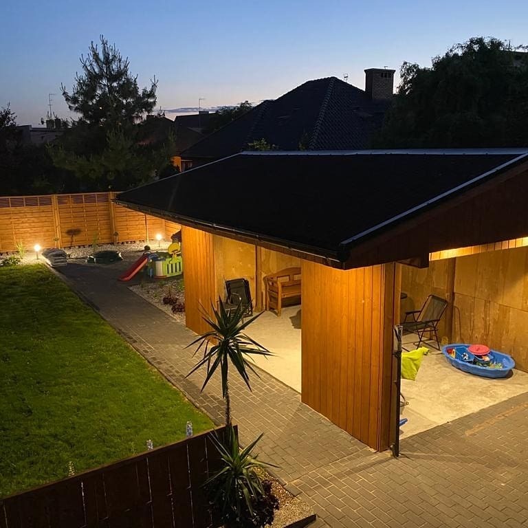 Oświetlony ogród z drewnianą altaną, placem zabaw i ścieżką z kostki brukowej. Lampy ogrodowe wzdłuż ścieżki. Widok na dom z ciemnym dachem. Wieczorny krajobraz.